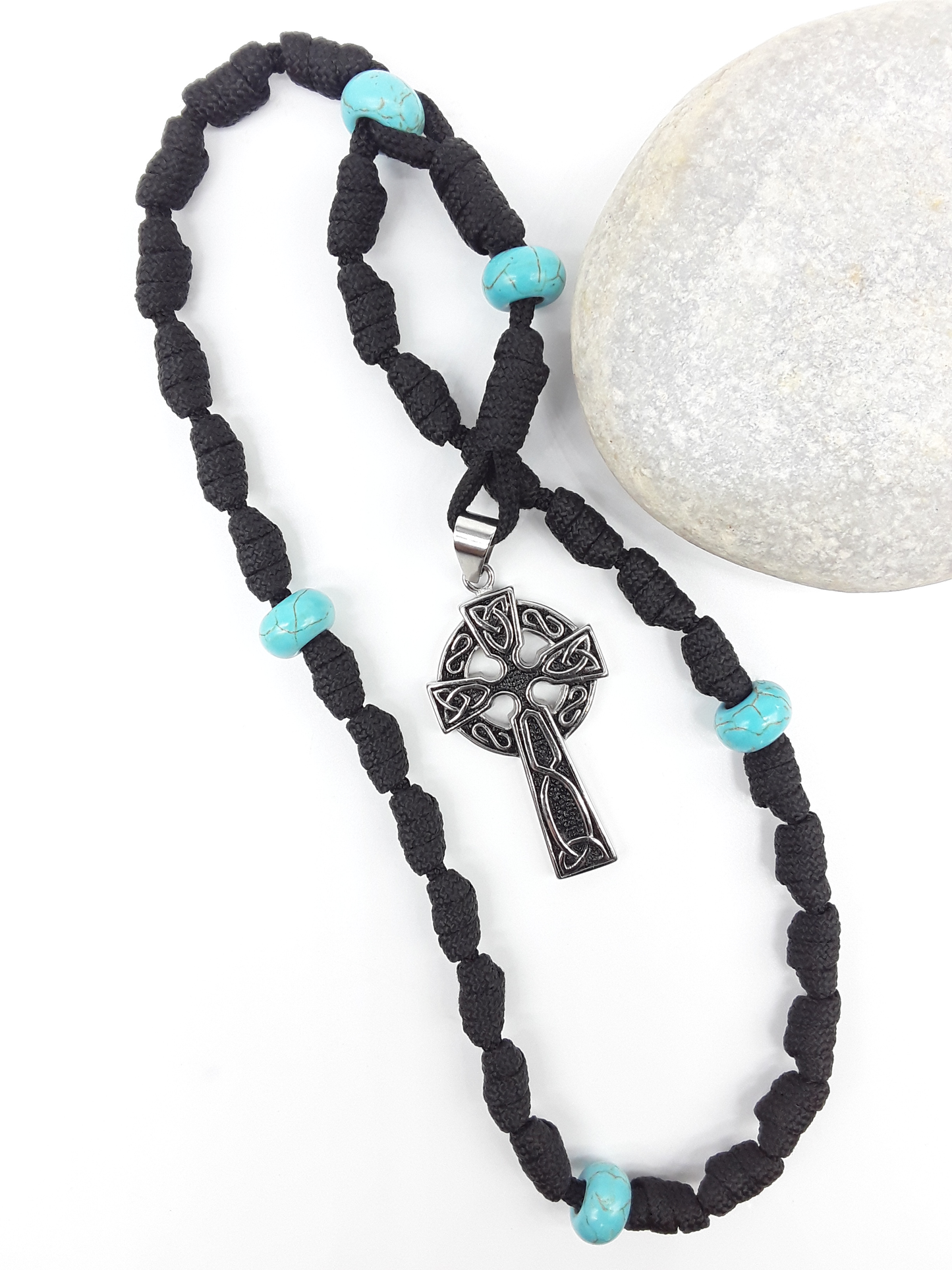 The Guiding Tide Celtic Trinity Anglican Rosary by Polmadeva with hand-knotted black paracord, Turquoise elements, and Celtic Trinity Cross beside stone.