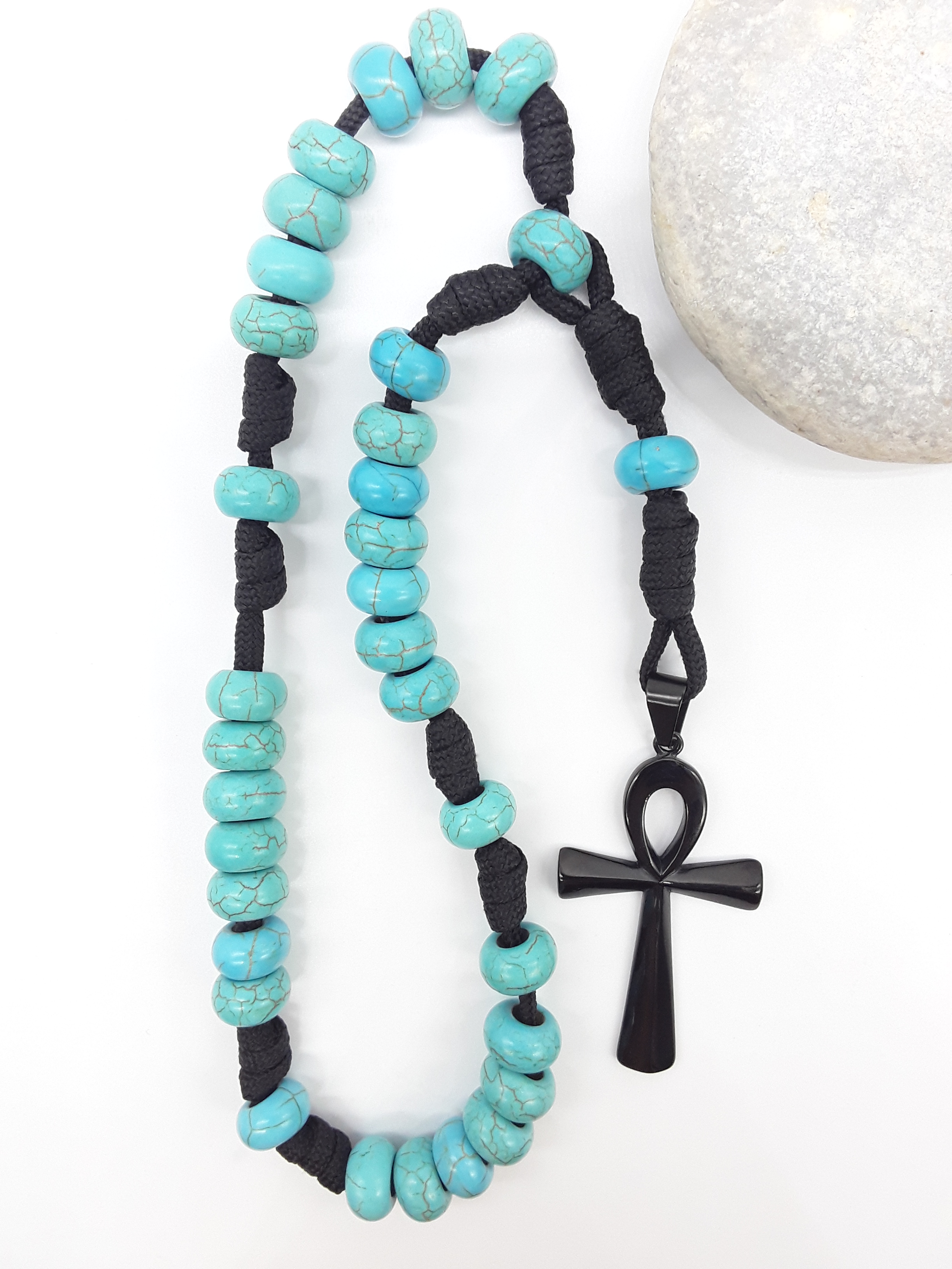 The Living Covenant Paracord Anglican Prayer Beads by Polmadeva beside natural stone, featuring Turquoise and Ankh cross