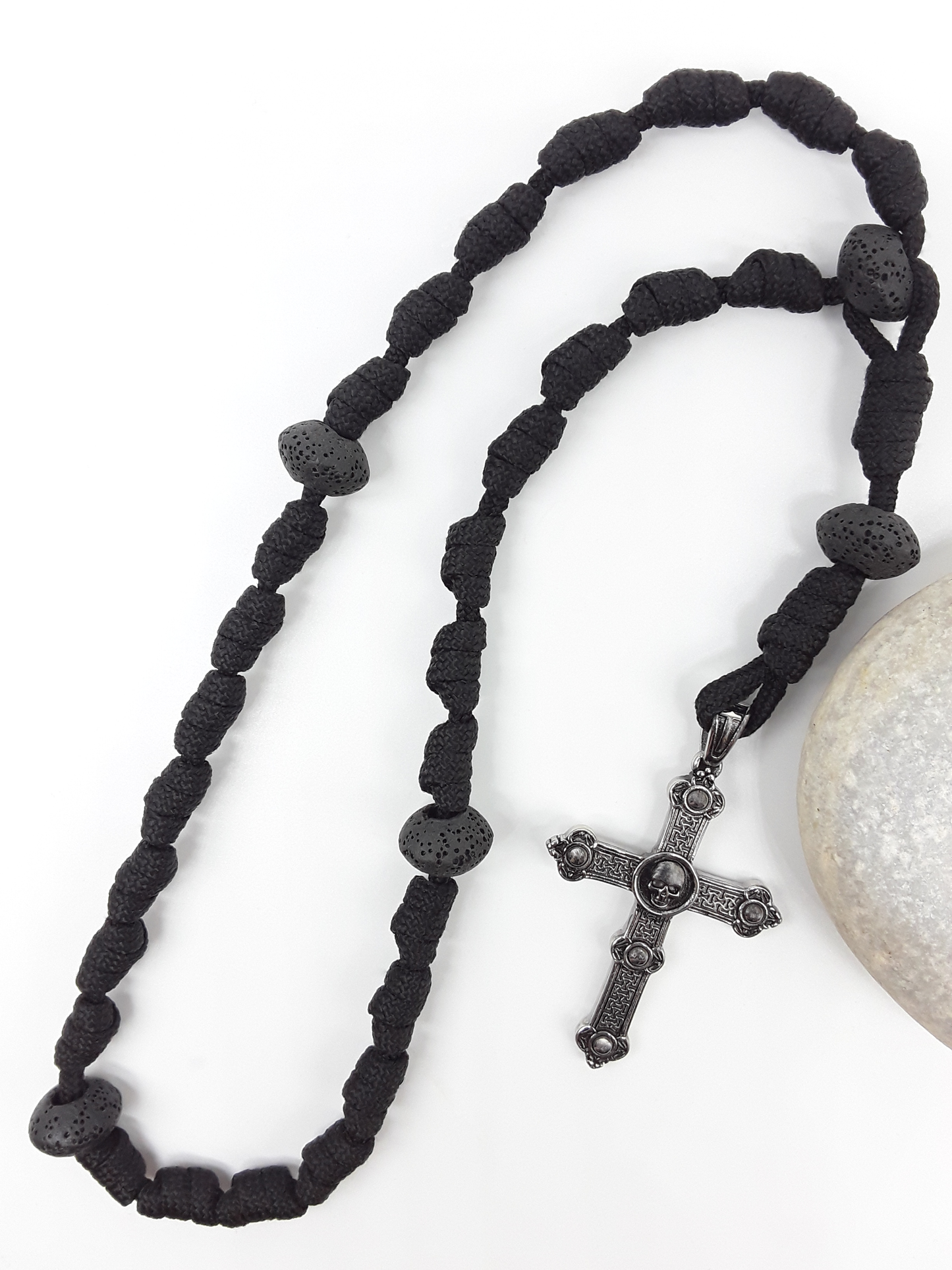 The Living Memory Paracord Anglican Prayer Beads by Polmadeva beside natural stone, featuring Volcanic Lava Stones and Skull Cross