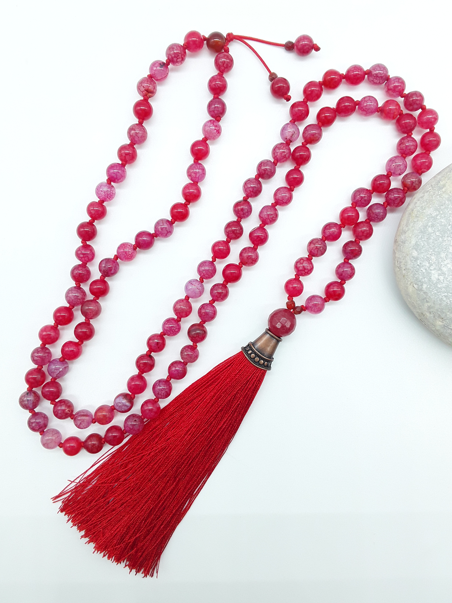 Dragon Veins Agate Mala Beads with red tassel placed beside white stone on white background, Polmadeva