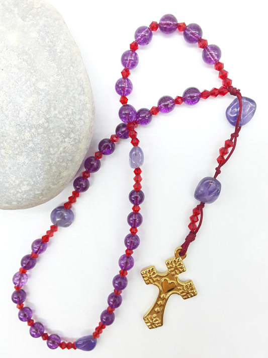 The Luminous Offering Anglican Prayer Beads by Polmadeva beside natural stone, featuring Amethyst, Purple Quartz, Swarovski crystals, and gold-tone cross