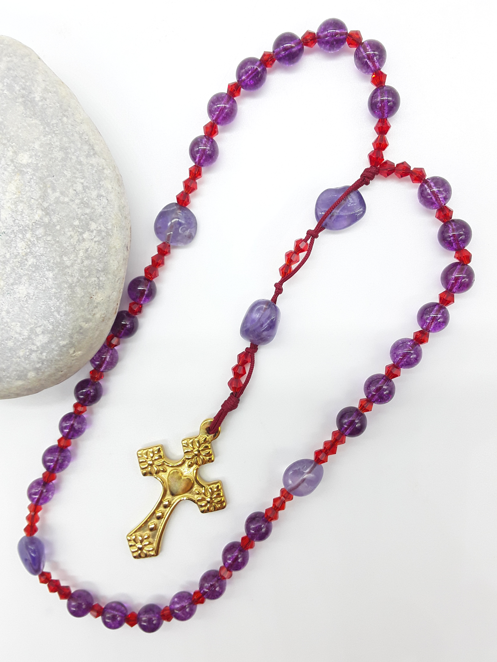 The Luminous Offering Anglican Prayer Beads by Polmadeva beside natural stone, featuring Amethyst, Purple Quartz, Swarovski crystals, and gold-tone cross