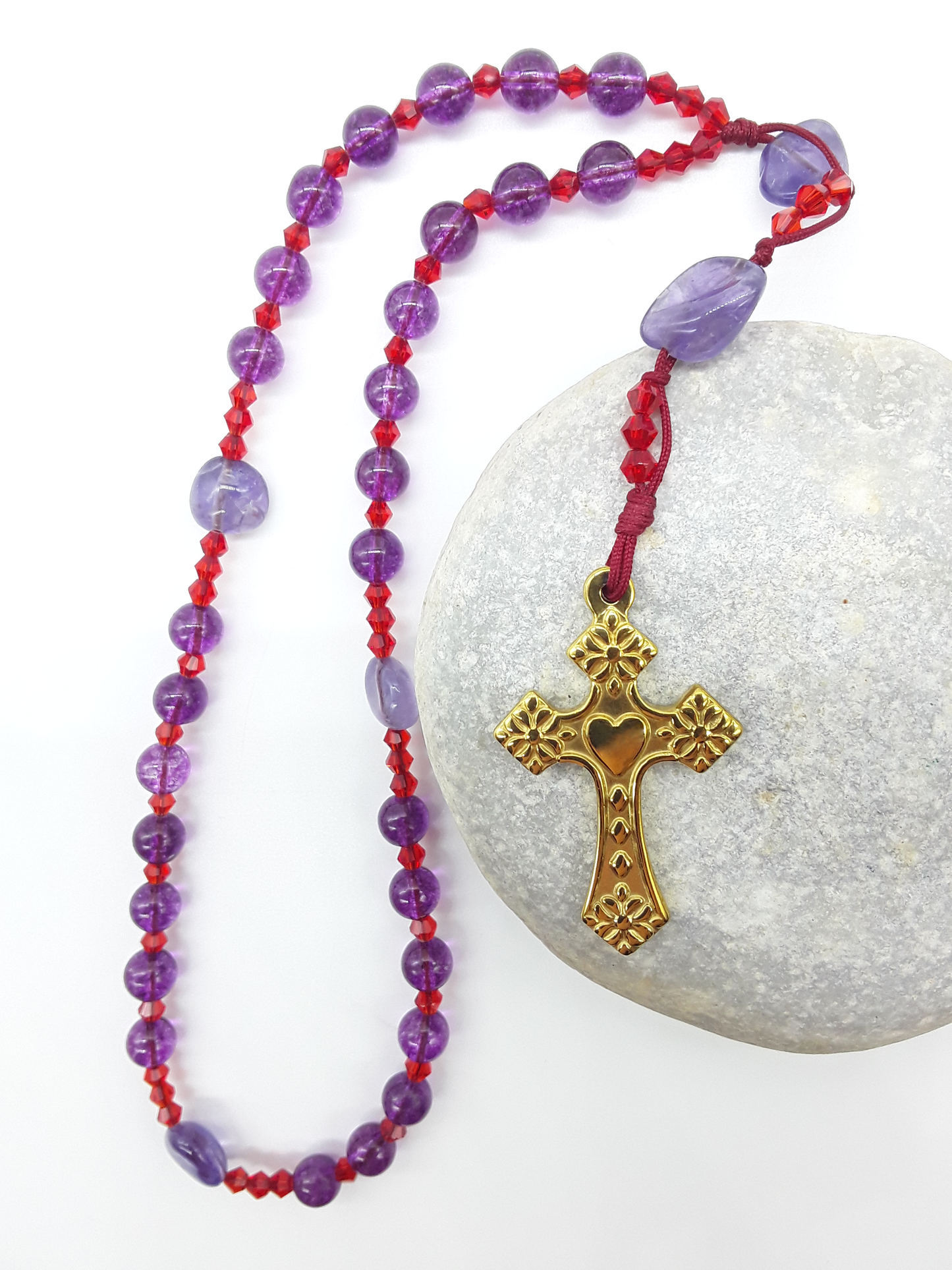 The Luminous Offering Anglican Prayer Beads by Polmadeva placed on stone, crafted with Amethyst, Purple Quartz, Swarovski crystals, and gold-tone cross