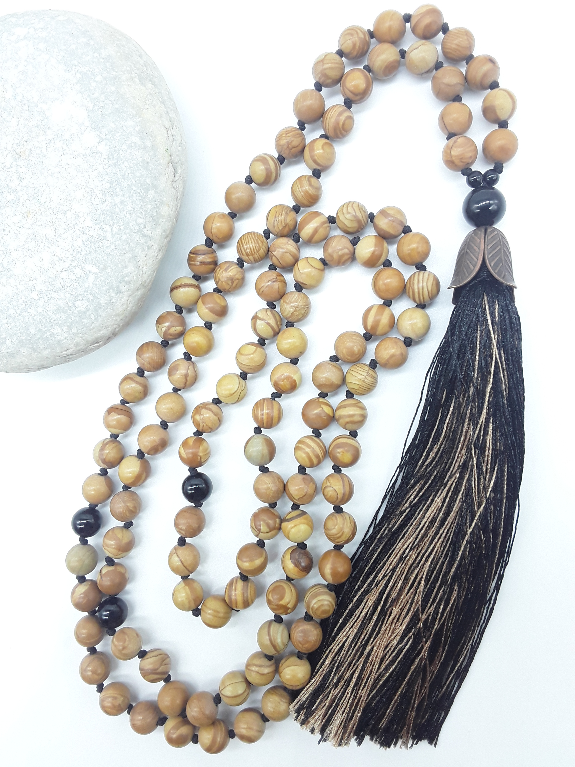 Tigerskin Jasper Mala Beads with Black Onyx details and tassel placed beside white stone on white background, Polmadeva