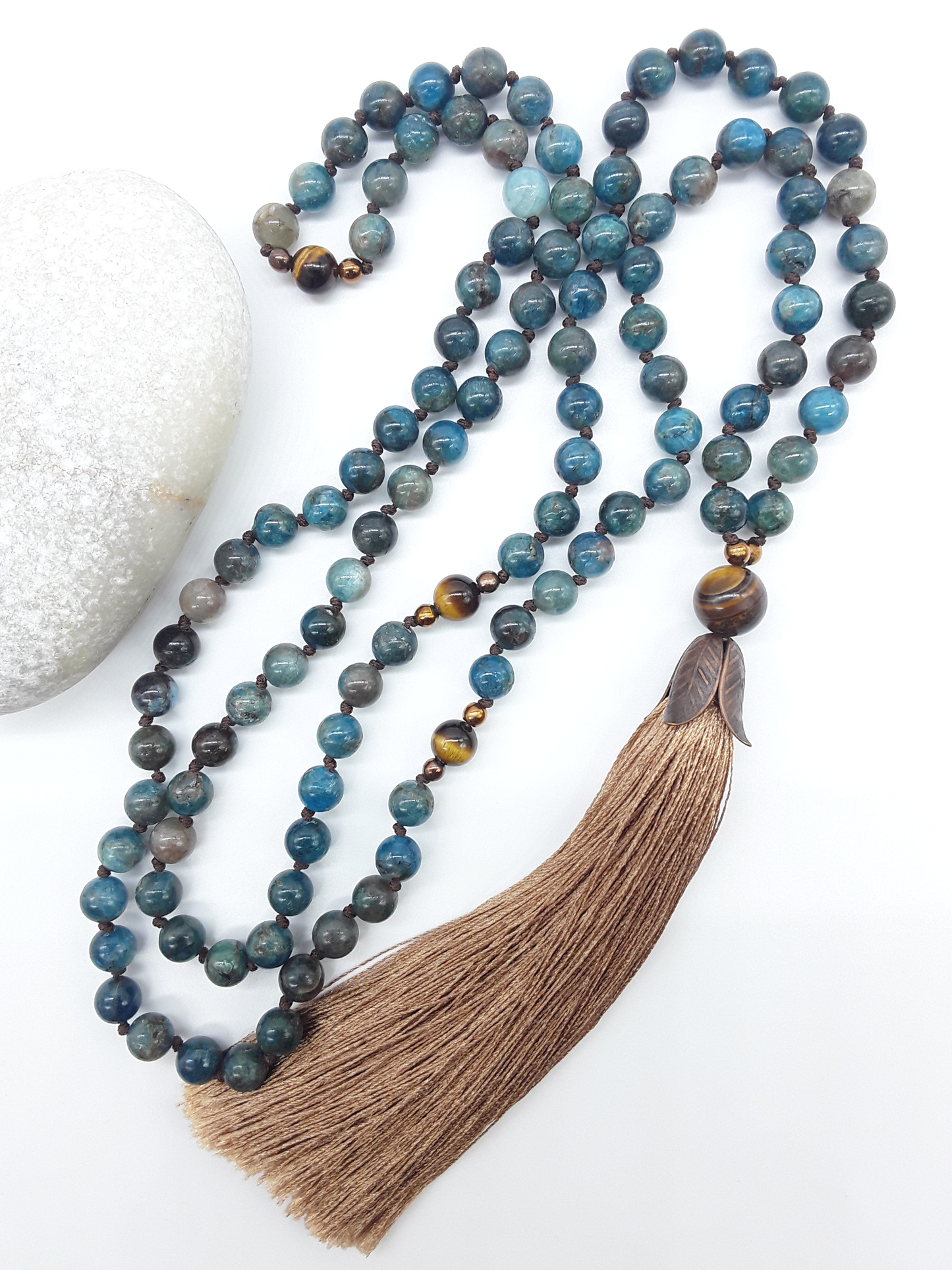 Apatite Mala Beads with Tiger’s Eye, Hematite details and tassel placed beside white stone on white background, Polmadeva