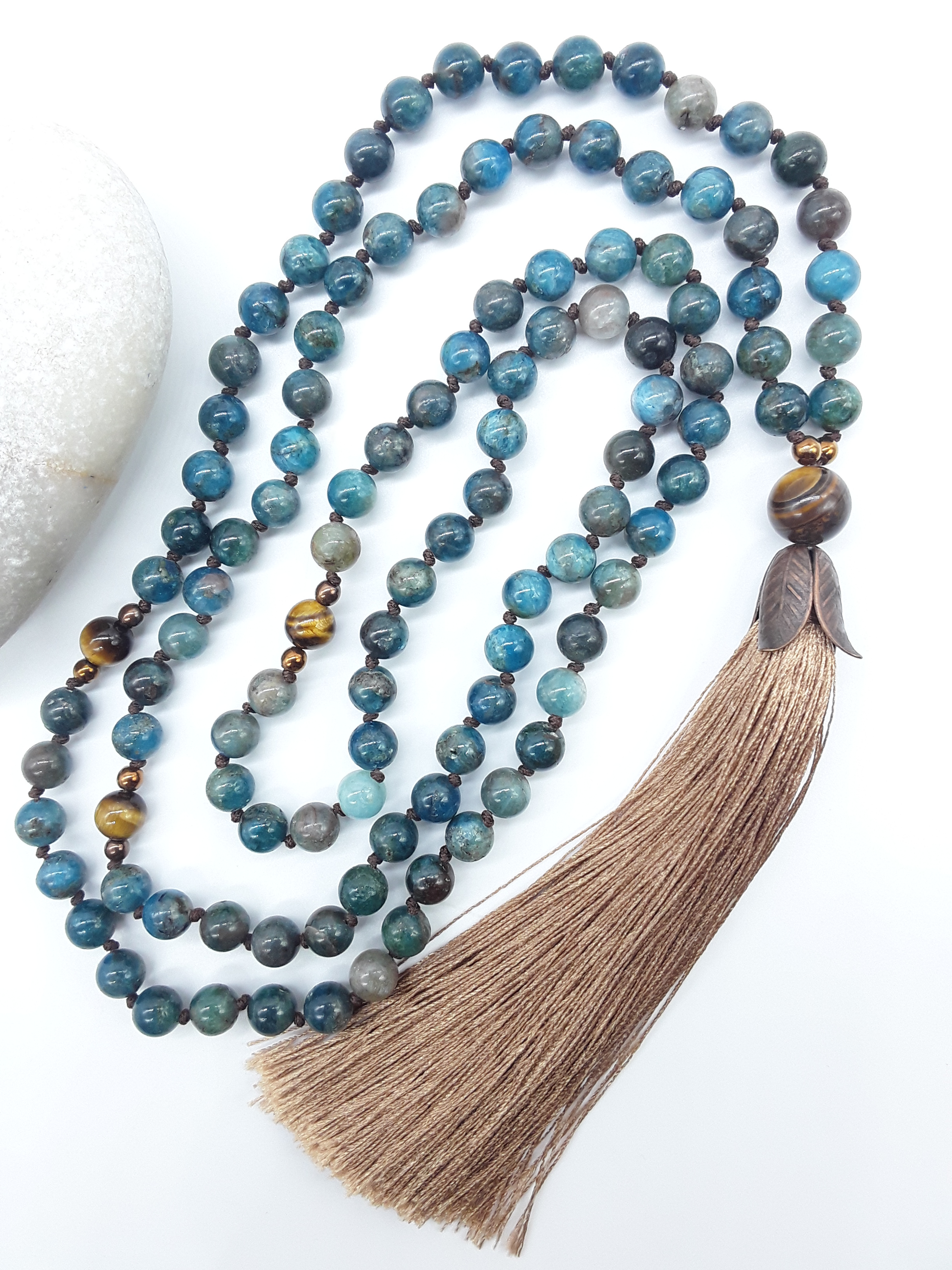 Apatite Mala Beads with Tiger’s Eye, Hematite details and tassel placed beside white stone on white background, Polmadeva