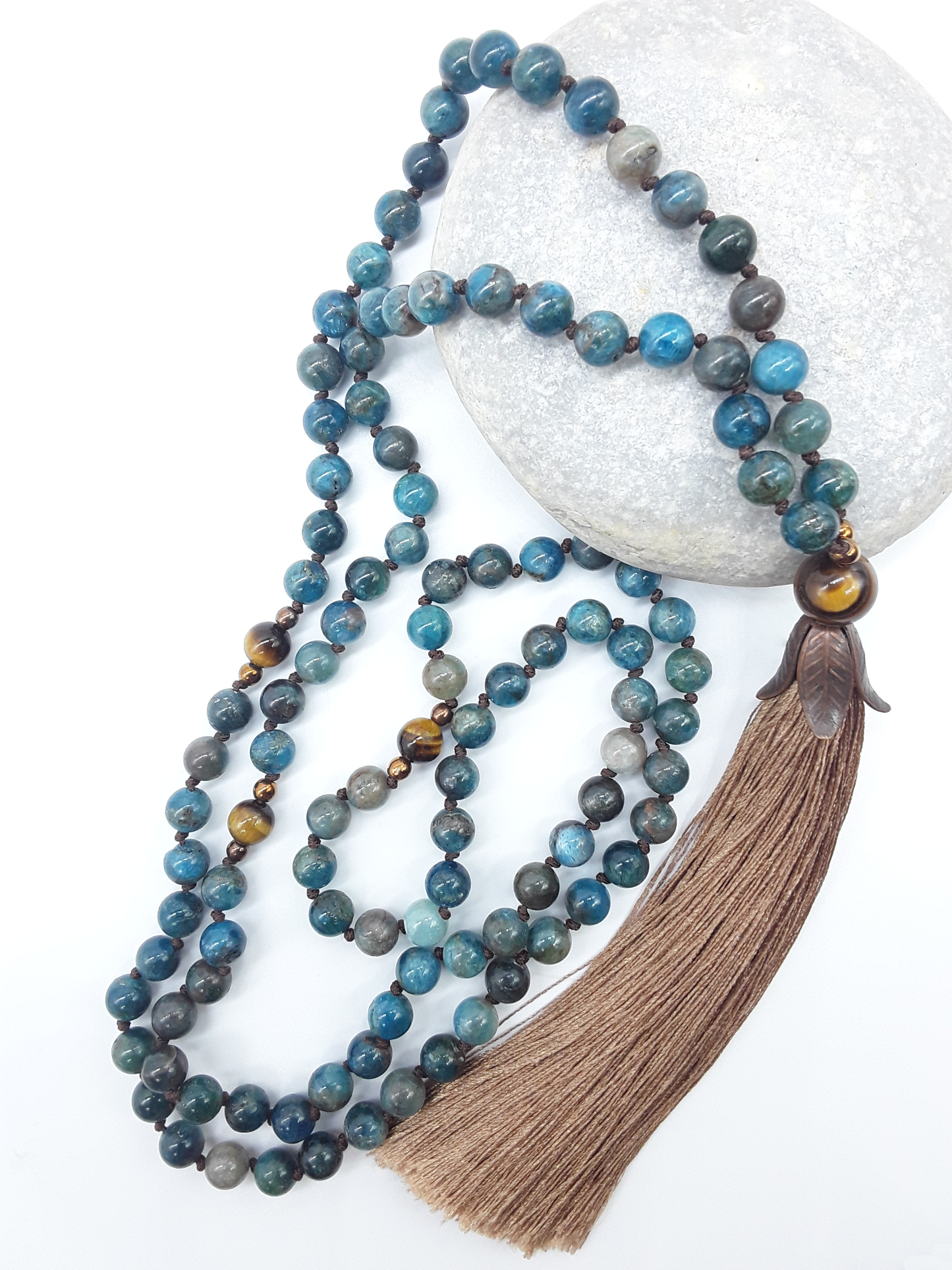 Apatite Mala Beads with Tiger’s Eye, Hematite details and tassel placed beside white stone on white background, Polmadeva