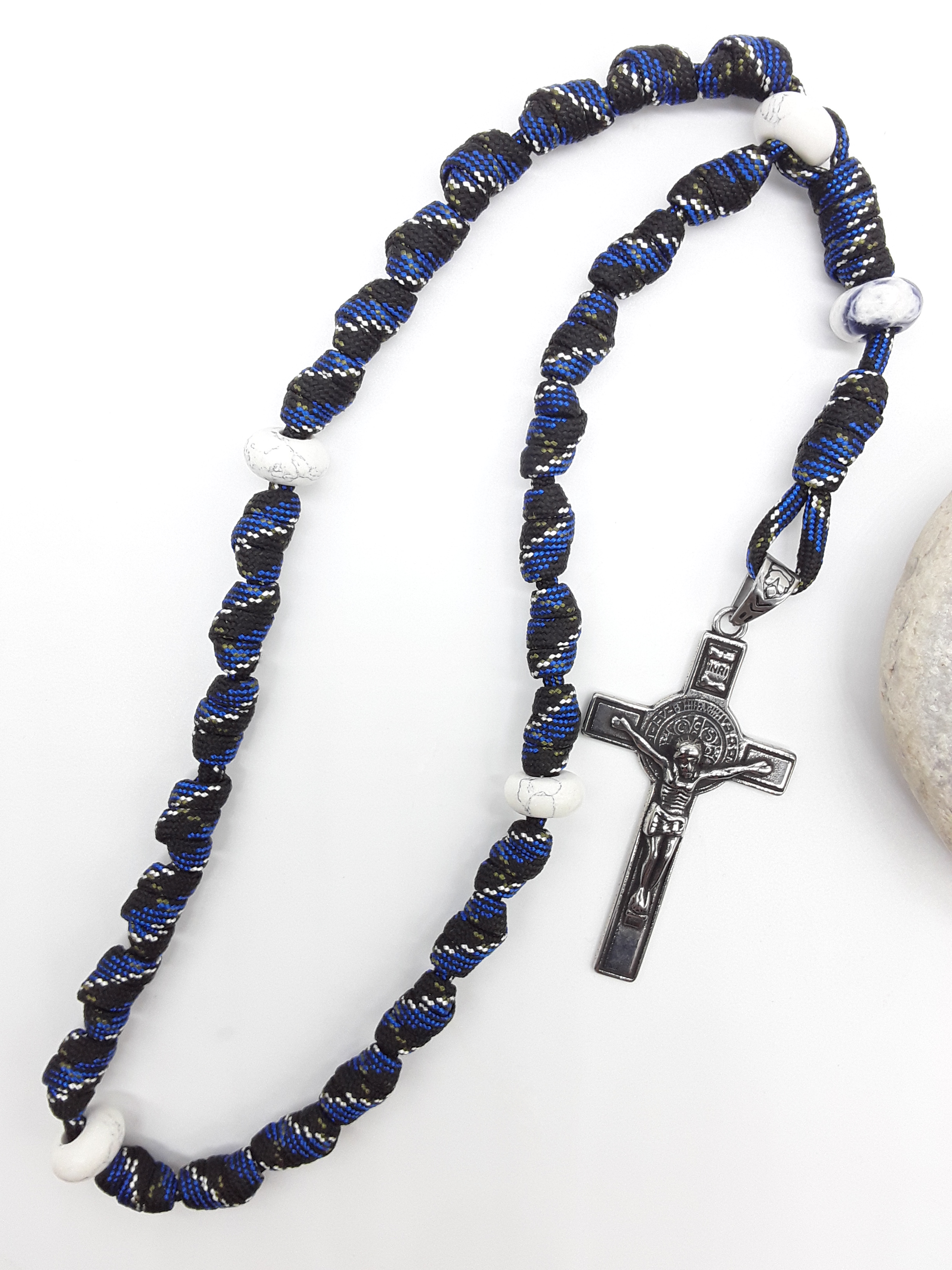 The Steady Witness Saint Benedict Anglican Rosary by Polmadeva with hand-knotted black and blue paracord, Howlite elements, and Saint Benedict Crucifix beside stone.