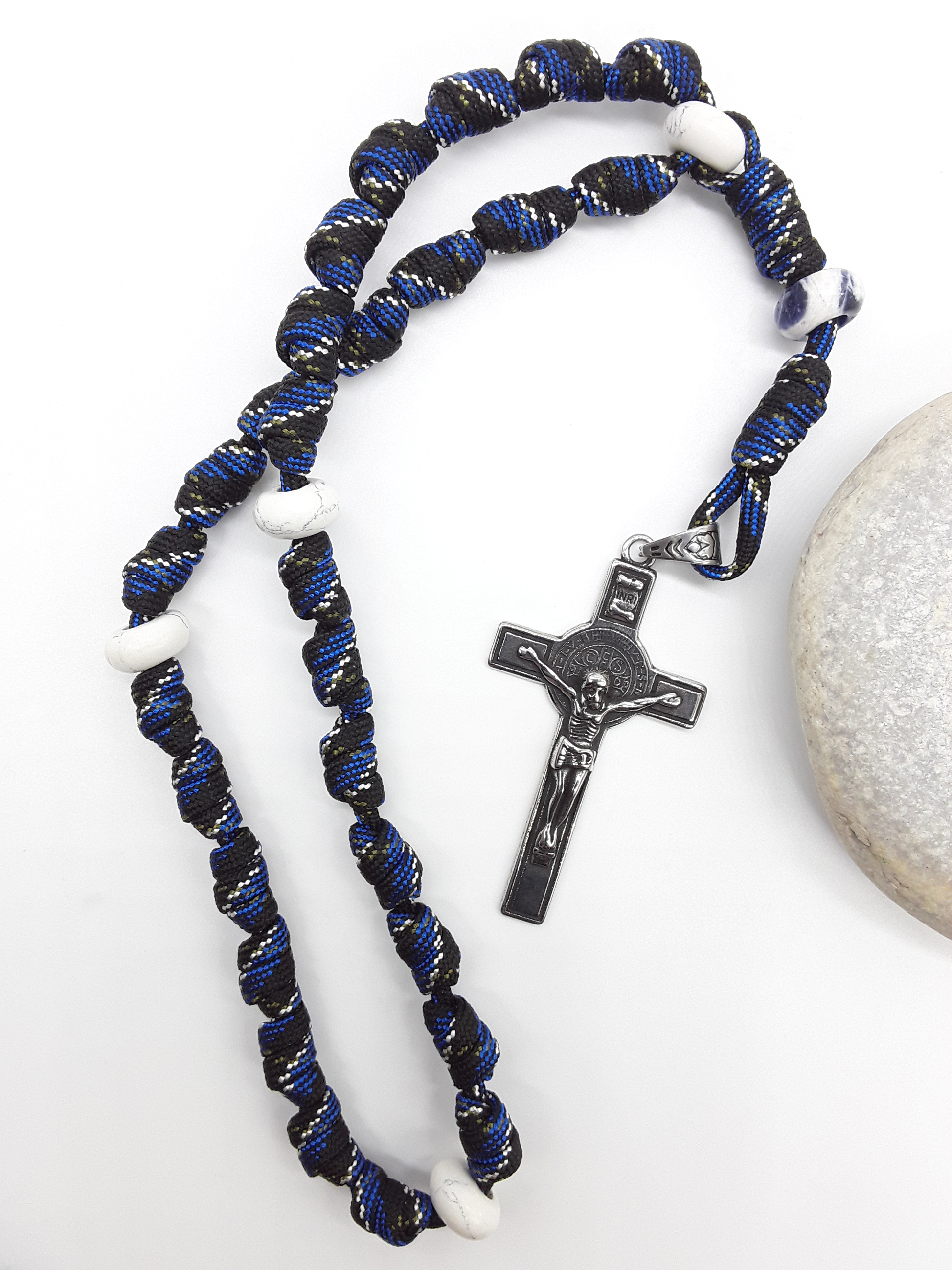 The Steady Witness Saint Benedict Anglican Rosary by Polmadeva with hand-knotted black and blue paracord, Howlite elements, and Saint Benedict Crucifix beside stone.