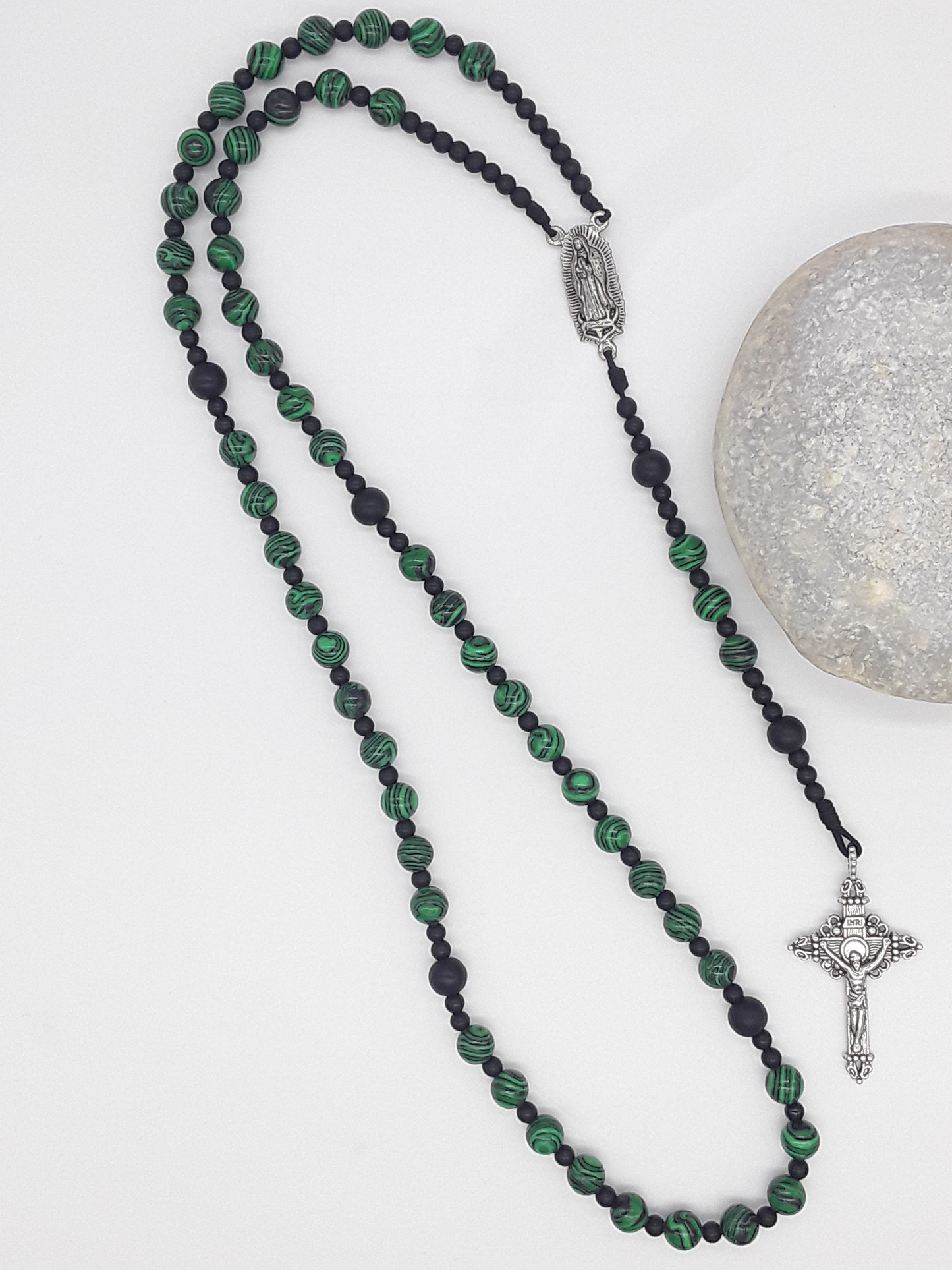 Catholic rosary made of black onyx and malachite beads with Our Lady of Guadalupe medallion and silver-toned crucifix, styled beside a natural stone.