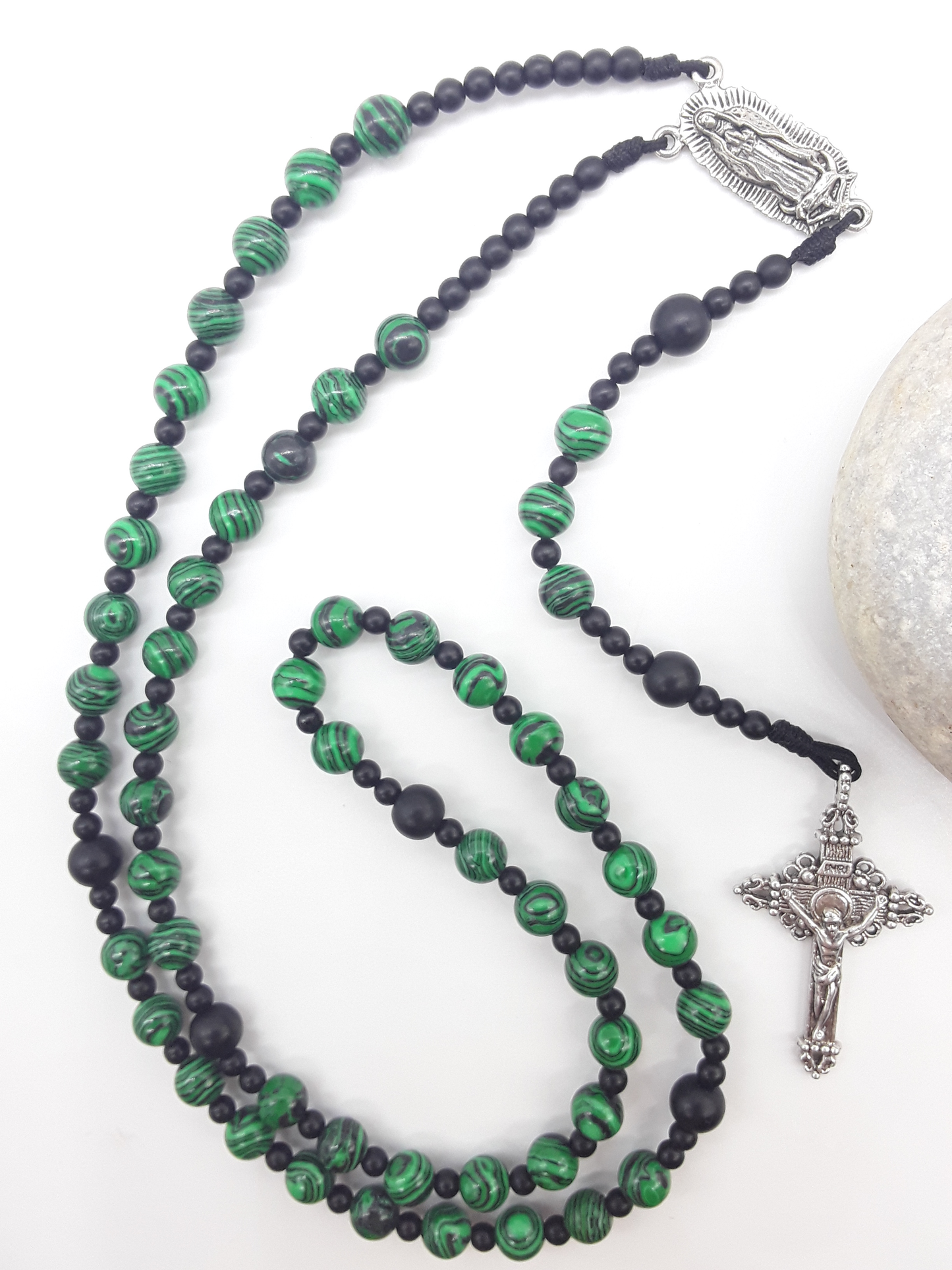 Catholic rosary made of black onyx and malachite beads with Our Lady of Guadalupe medallion and silver-toned crucifix, styled beside a natural stone.