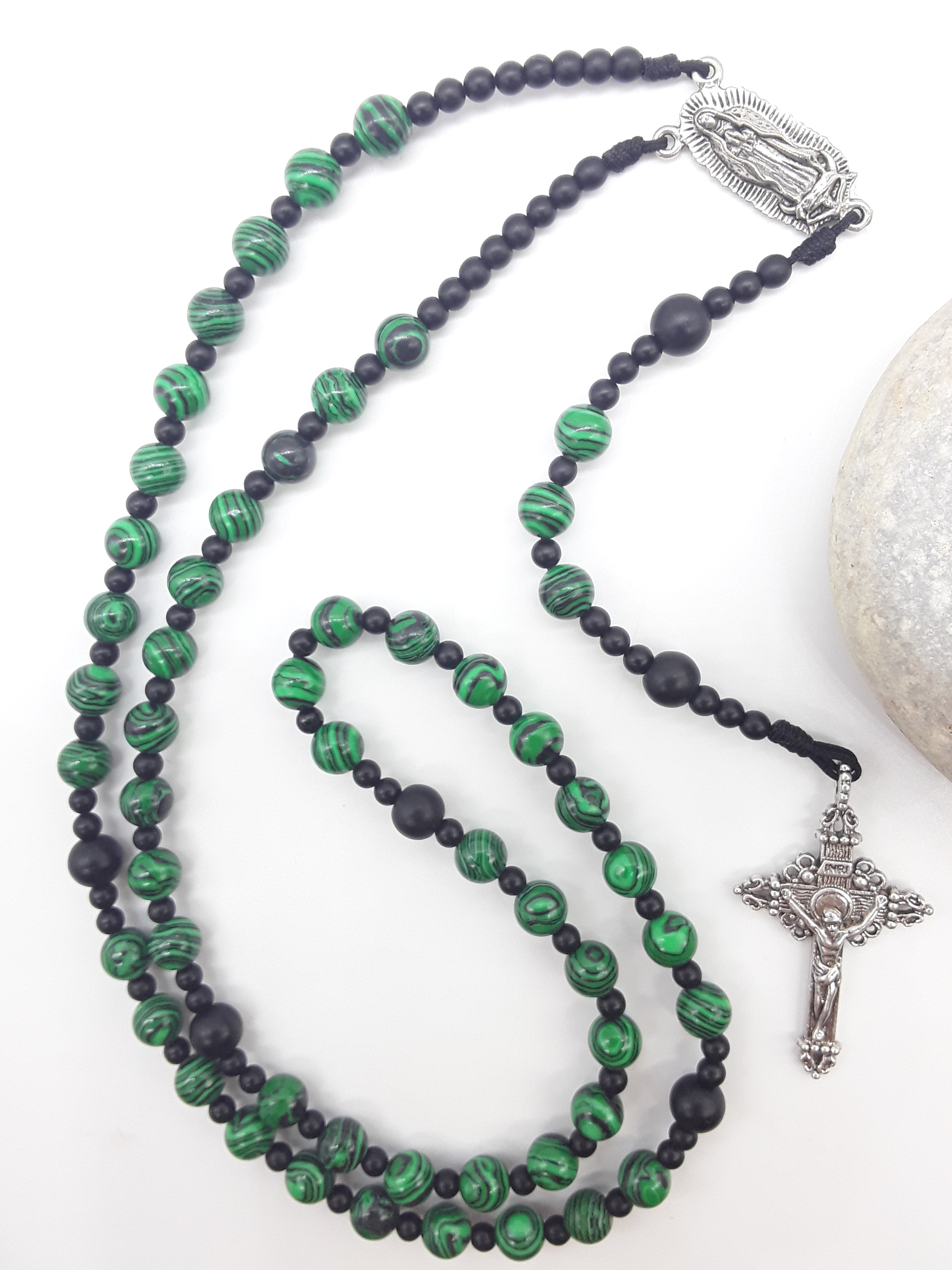 Catholic rosary made of black onyx and malachite beads with Our Lady of Guadalupe medallion and silver-toned crucifix, styled beside a natural stone.