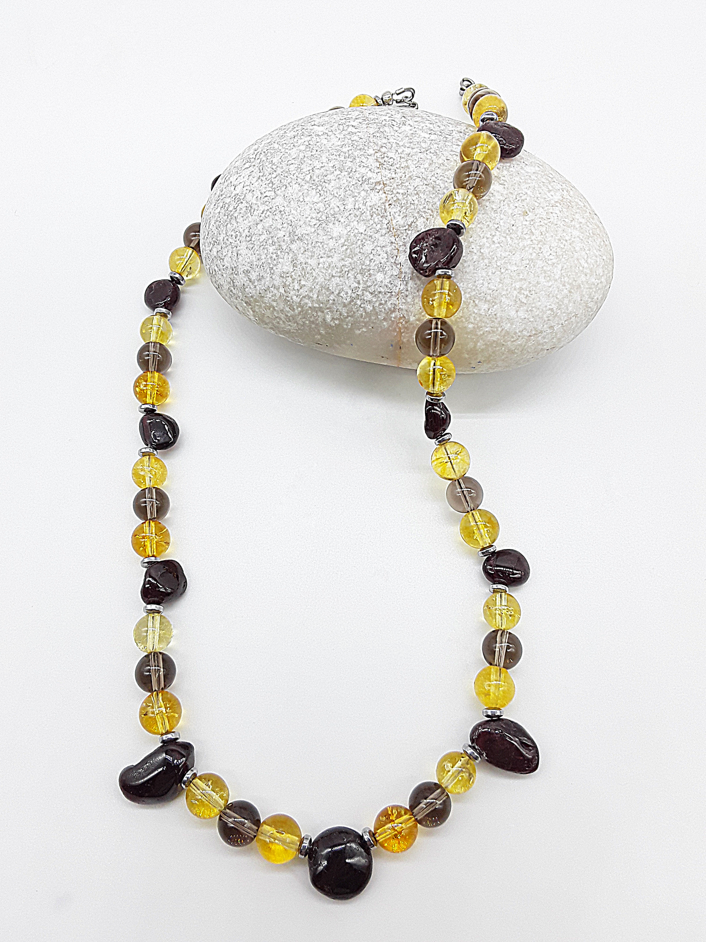 Gemstone necklace featuring citrine, garnet, smoked quartz, and hematite displayed beside a smooth white stone.