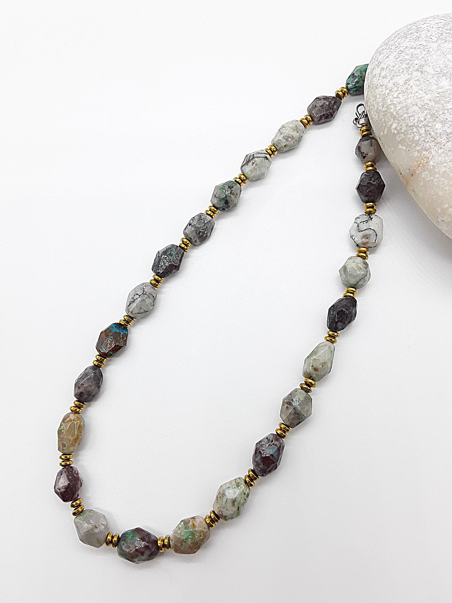 Natural stone necklace with multicolored faceted beads and gold spacers displayed on a white surface next to a smooth white stone.